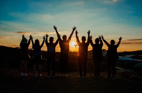 Gruppe von 7 Personen steht mit hochgehobenen Armen auf einem Hügel und sieht in den Sonnenuntergang - Reiseideen für Teams machen glücklich