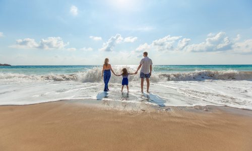 Servus Urlaub - Mutter, Tochter und Vater stehen Hand in Hand am Strand und eine Welle rollt heran